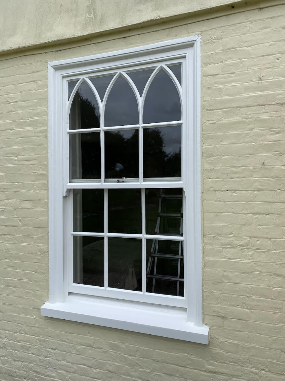 A closed white-framed window with an arched top section is set in a light yellow brick wall, featuring single glazed windows.