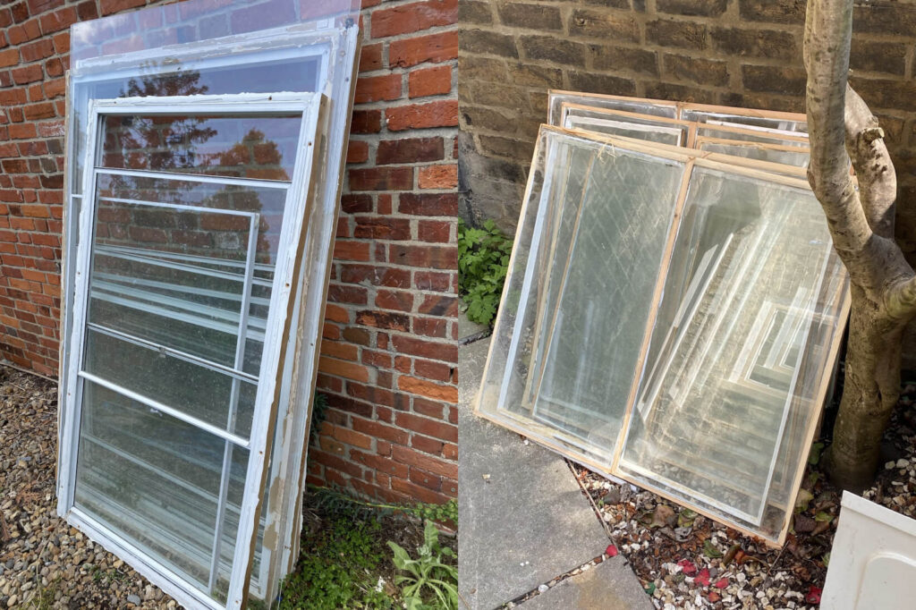 Two stacks of single glazing window frames lean against a brick wall and a stone wall, outdoors.