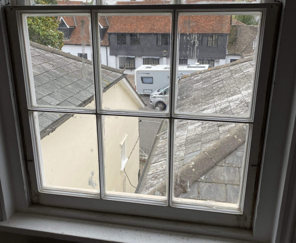 The view through Georgian sash reveals rooftops, a pristine white building, and a van parked on the quiet street below.