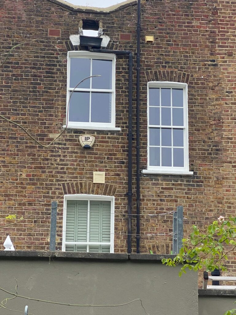 A brick building with three windows: one at the top with a broken arch, two on the middle floor, and one on the ground floor with closed shutters. A security alarm is mounted on the wall.