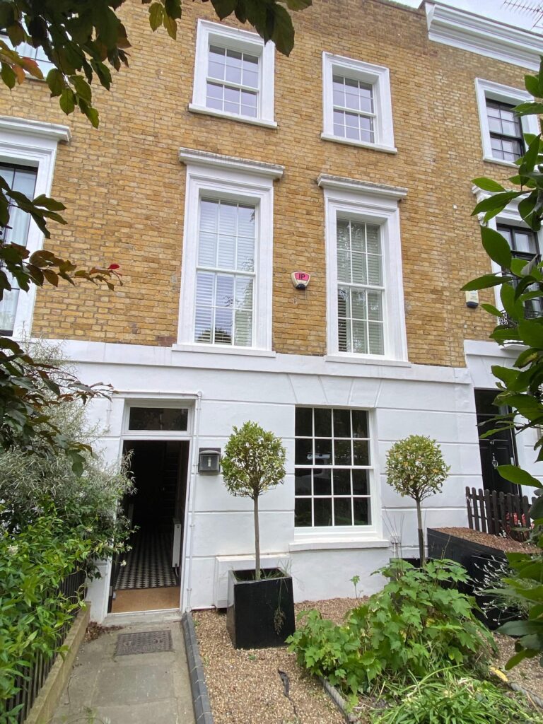 A brick and white-painted townhouse with three stories, large sash windows, a black door, potted trees, and a small front garden.