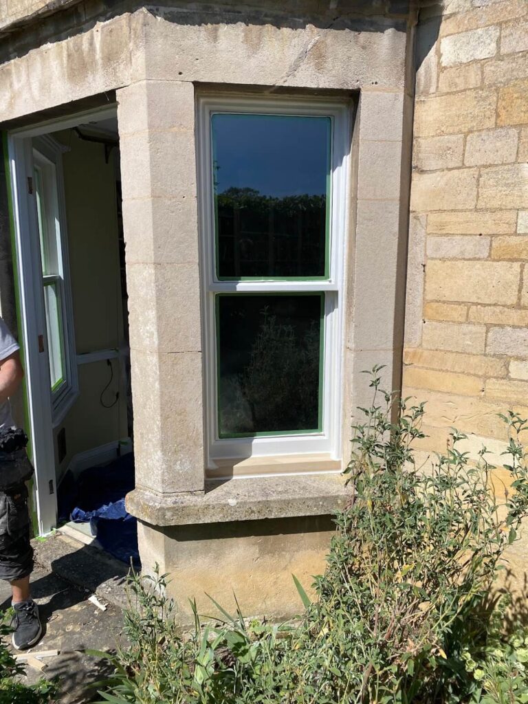 A recently installed white-framed sash window in a stone building, showcasing expert sash windows repair Leicestershire, with a partially open door and greenery in the foreground.