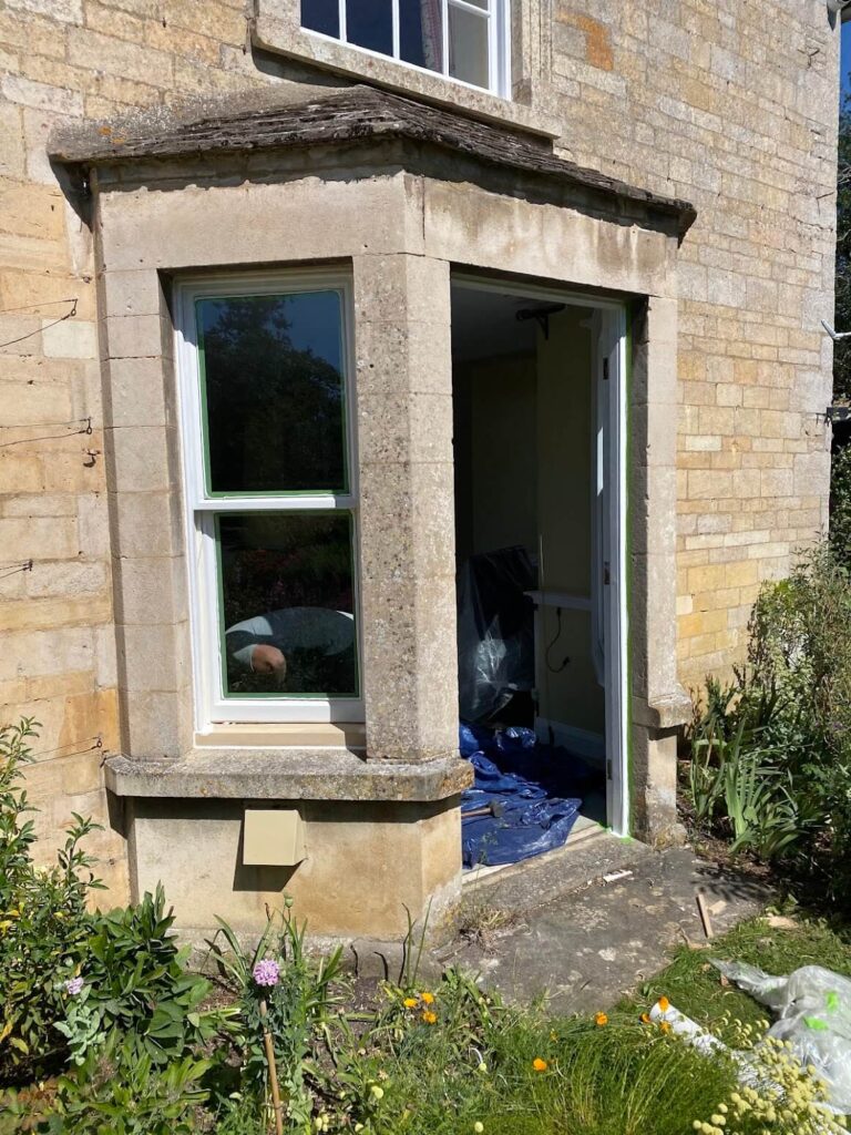 A stone house with a bay window under renovation; the door is open, protective sheets cover items inside, and garden plants bloom nearby. Sash windows repair Leicestershire services are underway to restore the home's charm.