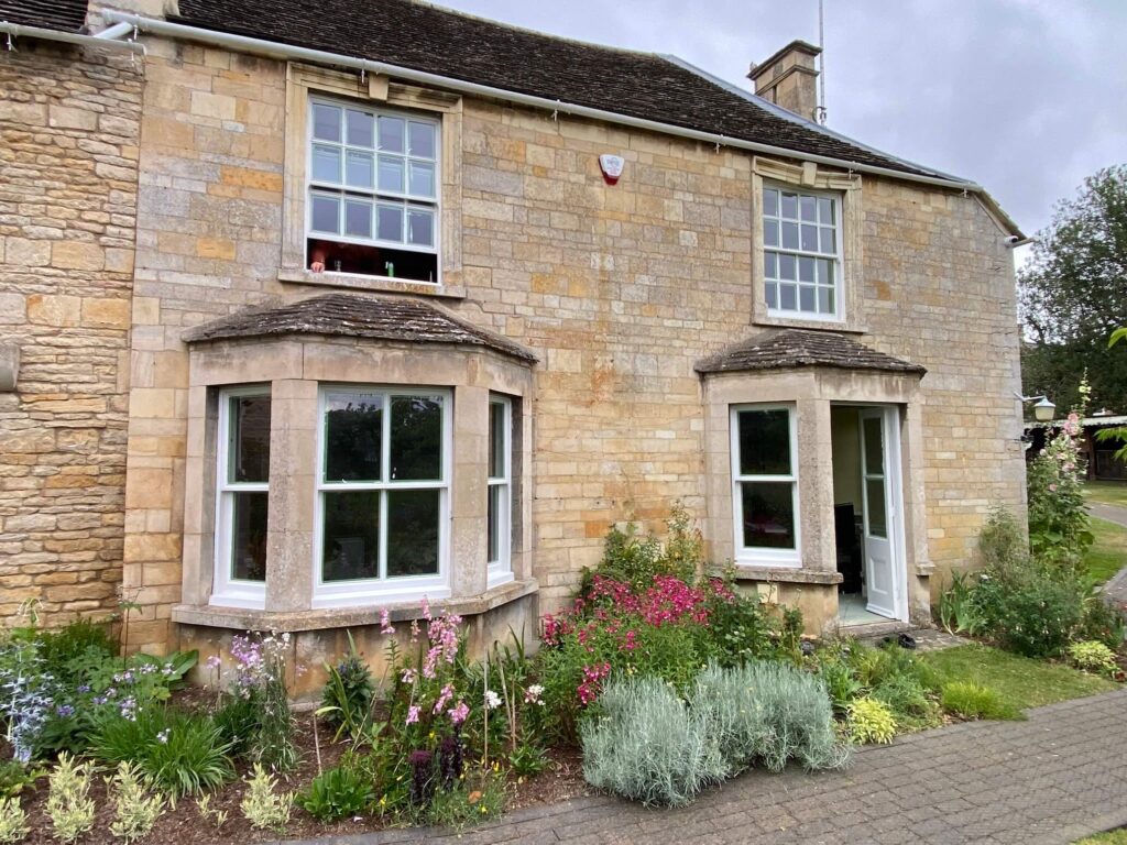 Stone house with two bay windows, a side door, and a small front garden with various flowering plants under a cloudy sky—ideal for those seeking sash windows repair Leicestershire services.