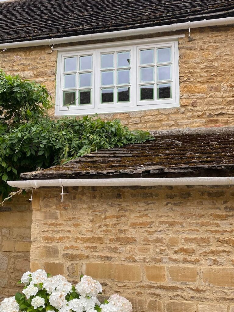 A stone house with a white-framed sash window, overgrown greenery on the roof, and white hydrangea flowers in the foreground—perfect inspiration for sash windows repair Leicestershire specialists.