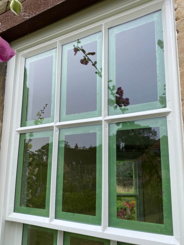 A window with six glass panes, each bordered with green painter's tape in preparation for painting—ideal for those seeking sash windows repair Leicestershire; garden and sky reflections shimmer in the glass.