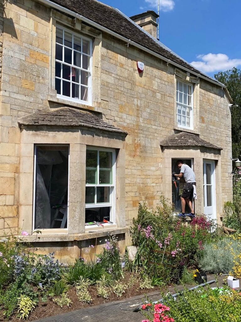 A person works on a window frame of a stone house with two bay windows, surrounded by a garden full of flowers—perfecting sash windows repair Leicestershire style amidst vibrant greenery.