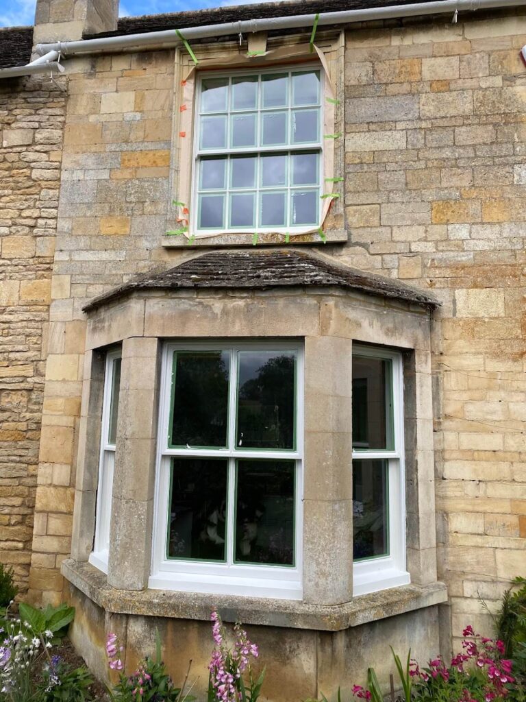 A stone house with a bay window on the ground floor and a rectangular window above, both with white frames; tape is visible around the upper window, suggesting recent sash windows repair Leicestershire.