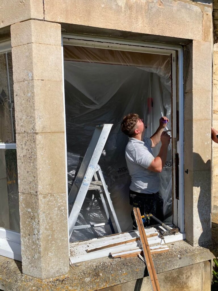 A person stands on a window ledge renovating a window frame, with plastic sheeting, a ladder, and tools visible—showcasing expert sash windows repair Leicestershire both inside and outside the opening.
