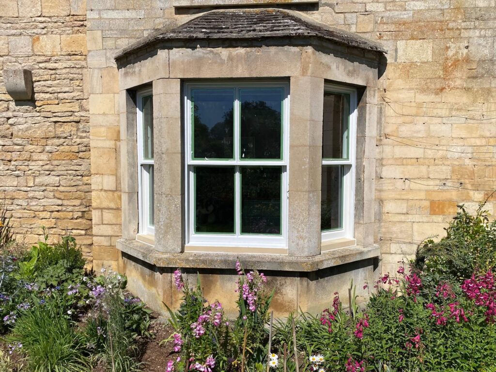 A stone bay window with white frames, possibly in need of sash windows repair Leicestershire, set in a brick wall and surrounded by a garden with blooming flowers.