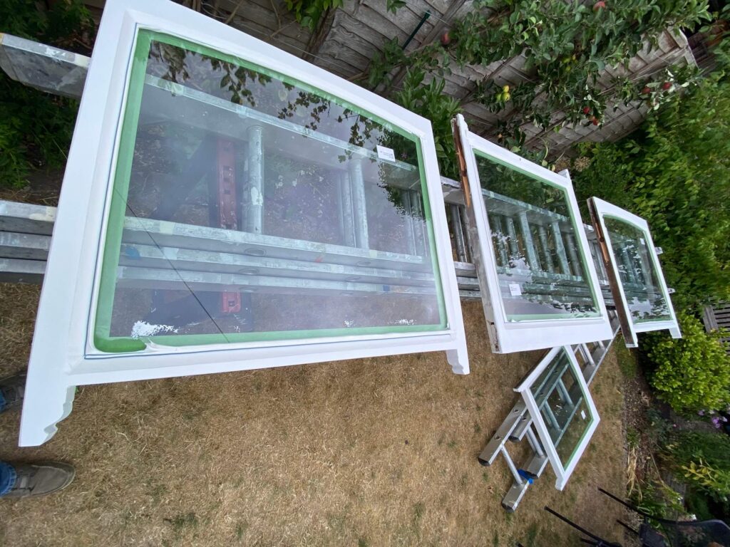 Several white-framed glass windows are laid out on ladders and stands in a garden, likely being prepared or painted as part of sash windows repair Leicestershire. Shrubs and a wooden fence are visible in the background.