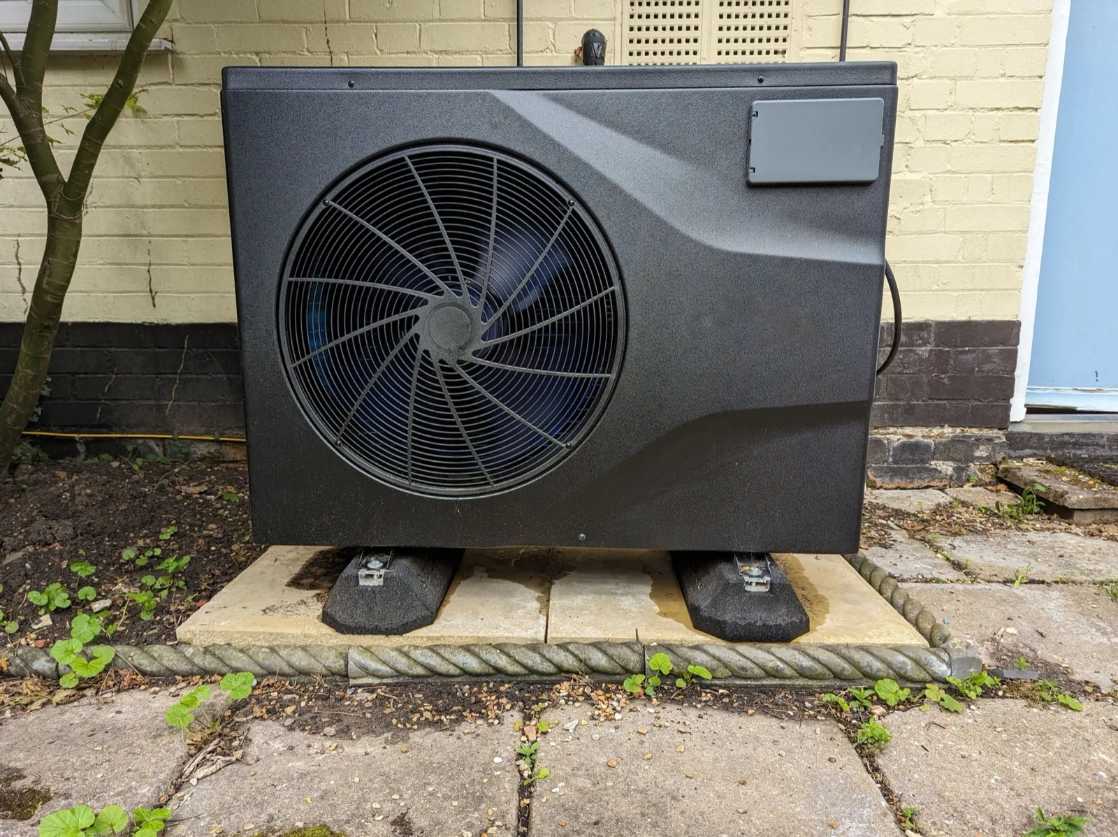 A dark air source heat pump unit on a paved base outside an older house with cream-painted brick walls