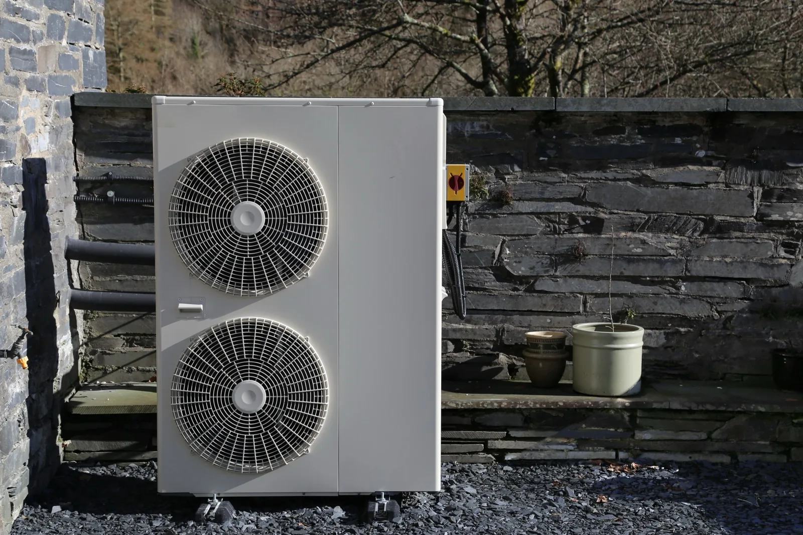 A twin-fan air source heat pump standing on a gravel bed beside a slate stone garden wall