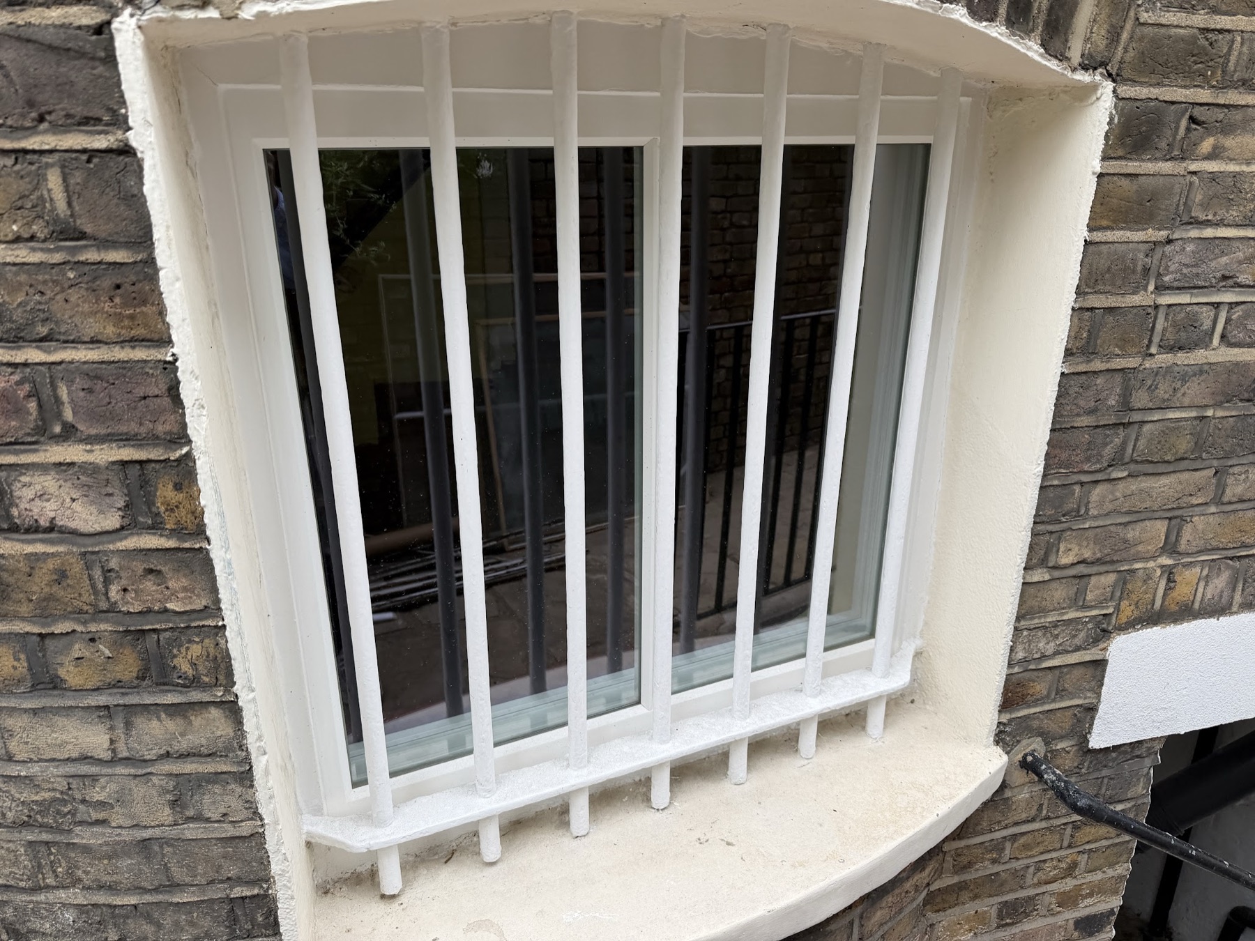 Small lower-ground window with a shallow curved head reglazed with a vacuum unit behind white-painted security bars